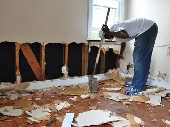 A worker removing wet drywall from the lower walls of a room after flooding